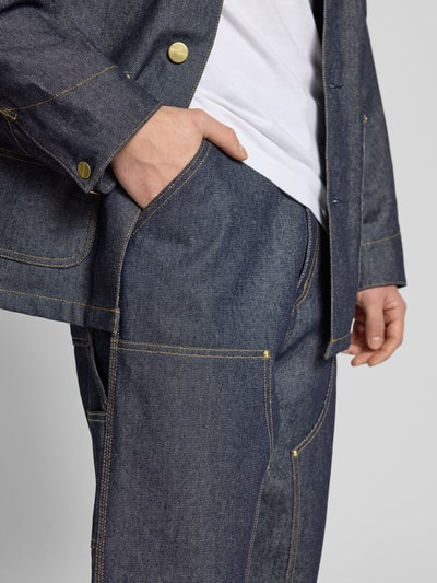 Männermodel trägt Jeans mit Eingrifftaschen von Carhartt Work In Progress in Dunkelblau, grobe Struktur, Kontrastnähte, Nahaufnahme Oberkörper.