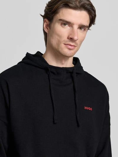 Męski model nosi Relaxed Fit Hoodie aus Baumwoll-Mix od HUGO, jednokolorowy, wyhaftowane logo, mieszanka bawełny, faktura wafla, długi rękaw, kolor czarny, zdjęcie: zbliżenie na górę ciała.