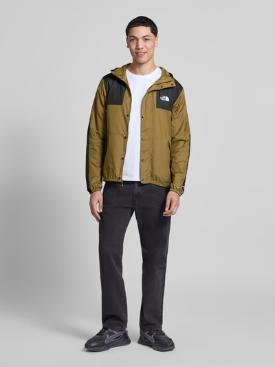 Mannelijk model draagt Regular Fit Jacke mit Logo-Print model 'Mountain' van The North Face in olijfgroen met zwarte details, steekzakken met ritssluiting, elastische boorden, capuchon met tunnelkoord, ganzekörperaanzicht van voren, ruglengte maat M 72 cm, schouderbreedte 48 cm, borstwijdte 53 cm, mouwlengte 70 cm.