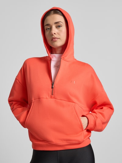 Modelka kobieca nosi Hoodie mit Reißverschluss und Label-Stitching od Under Armour, bawełniana mieszanka, krój regular fit, zamek, przeszycie z logo, kieszeń kangurka, koralowy kolor, gładka tekstura, widok na górną część ciała z przodu