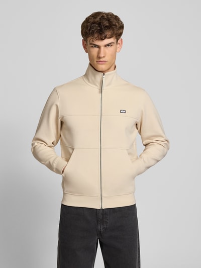 Männermodel trägt Sweatjacke mit Label-Patch Modell 'KOSTER' von Helly Hansen in Beige, Baumwoll-Mix mit Stretch-Anteil, Gerader-Schnitt, Stehkragen, Reißverschluss, Seitliche Eingrifftaschen, Rückenlänge bei Größe M 67 cm, Ärmellänge 66 cm; Ganzkörperansicht von vorne.