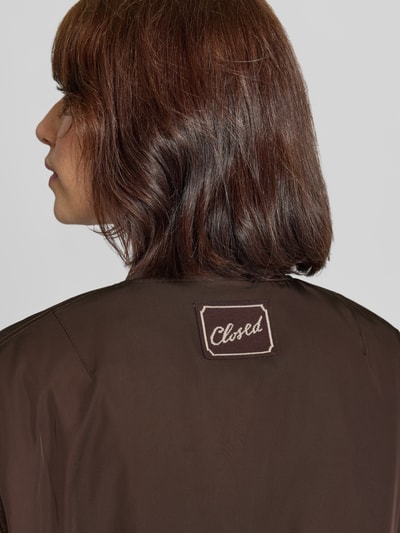 Frauenmodel trägt Jacke mit Stehkragen und verdecktem Reißverschluss von Closed in Dunkelbraun, glatte Struktur, Detailaufnahme Rückansicht, Relaxed Fit, sichtbares Markenlabel, Kontext: Pattentaschen, Rückenlänge 60 cm, Schulterbreite 46 cm, Brustweite 56 cm, Ärmellänge 60 cm.