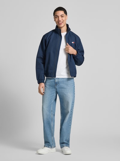 Męski model nosi Regular Fit Blouson mit Stehkragen od Tommy Jeans, efekt: jednolity kolor, krój regular fit, stójka, długi rękaw, wpuszczane kieszenie, kieszeń wewnętrzna, naszywka z logo, zapięcie kryty zamek błyskawiczny, elastyczne wykończenie, widok całej sylwetki z przodu.