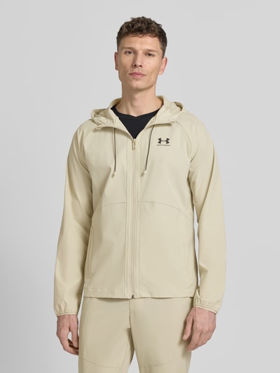 Męski model nosi Regular Fit Jacke mit Logo-Print Modell 'Vibe' od Under Armour, elastyczny materiał, kaptur, zamek, jasny beż, krój regular fit, długość tyłu 70 cm, długość rękawa 72 cm, widok całej sylwetki z przodu.