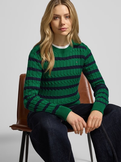 Frauenmodel trägt Slim Fit Strickpullover aus reiner Baumwolle von Tommy Hilfiger in grün-blauem Streifenmuster, Rundhals, Rippenabschlüsse, Logo-Detail, Nahaufnahme Oberkörper.