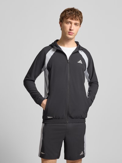 Männermodel trägt Sweatjacke mit Kapuze von adidas Training in Schwarz mit grauen Details, Material-Mix mit Stretch-Anteil, Lockerer Schnitt, Reißverschluss, Label-Print und Eingrifftaschen, Ganzkörperansicht von vorne.