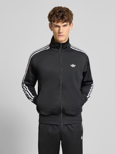 Männermodel trägt schwarze Sweatjacke mit Reißverschluss von adidas Originals, weiße Streifen, Stehkragen, Regular Fit, Ganzkörperansicht von vorne.