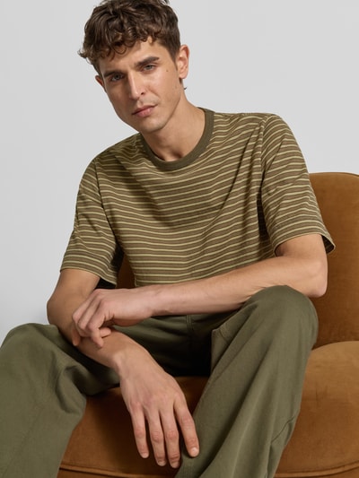 Männermodel trägt Relaxed Fit T-Shirt mit Label-Stitching von Forét, reine Baumwolle, Olivgrün mit feinen Streifen, Halbarm, Nahaufnahme Oberkörper.