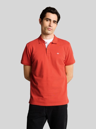 Männermodel trägt Regular Fit Poloshirt aus reiner Baumwolle van Tom Tailor, effen rood, korte mouw, piqué structuur, logo-stiksel, polokraag, 2-knoopssluiting, split aan zijkant, vooraanzicht van bovenlichaam.