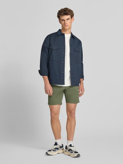 Männermodel trägt Straight Fit Shorts mit abnehmbarem Gürtel Modell 'Adam' von Jack & Jones aus Baumwoll-Mix in Olivgrün, Ganzkörperansicht von vorne. Shorts mit Eingriff- und Gesäßtaschen, Knopf- und Reißverschluss, Bundweite 40 cm, Innenbeinlänge 19 cm, geeignet als Herren Shorts.