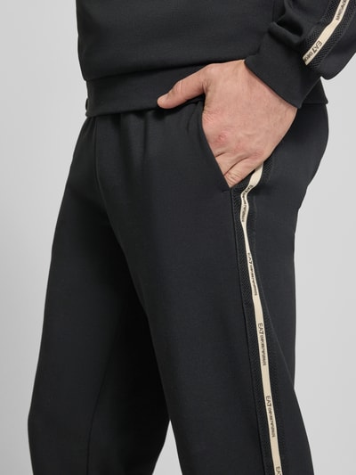 Männermodel trägt schwarze Sweatpants aus Baumwoll-Mix von EA7 Emporio Armani, beige Seitenstreifen, Eingrifftaschen, Nahaufnahme Oberkörper.