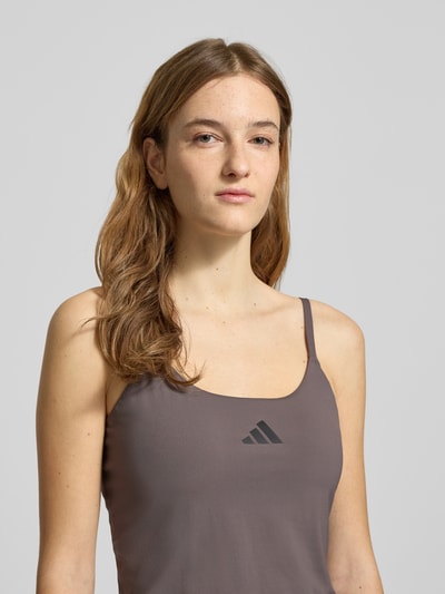 Modelka kobieca nosi Tanktop mit Label-Print od adidas Sportswear w kolorze szarobrązowym, materiał z domieszką stretchu, krój slim fit, nadruk z logo, długość tyłu w rozmiarze S: 26 cm, zbliżenie na górną część ciała, widok z przodu.