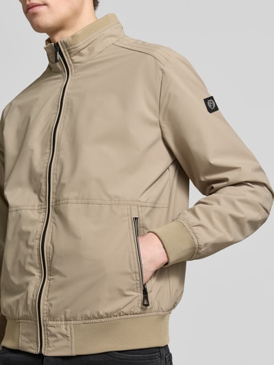 Męski model nosi Regular Fit Jacke mit Stehkragen od Lerros, beżowa kurtka z elastycznego materiału, logo, zamek, kieszenie, krój regular fit, długość tyłu w rozmiarze M 67 cm, szerokość ramion w rozmiarze M 49 cm, szerokość w rozmiarze M 58 cm, długość rękawa w rozmiarze M 68 cm, detalająca zbliżenie na górną część ciała z przodu