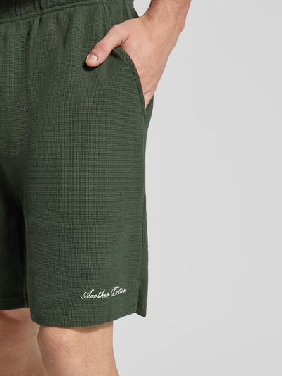 Männermodel trägt Shorts mit Logo-Print und Eingrifftaschen von ANOTHER COTTON LAB, dunkelgrün, Baumwoll-Mix, Nahaufnahme Oberschenkel.