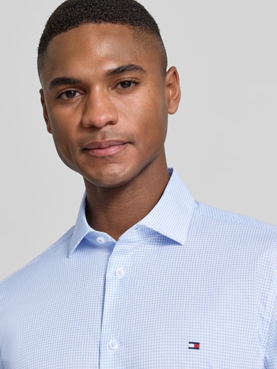 Mężczyzna model nosi Slim Fit Freizeithemd aus Baumwoll-Mix von Tommy Hilfiger Tailored, jasnoniebieskie, lekko elastyczne, wzór w drobną kratkę, logo na piersi, materiał bawełniany, krój slim fit, zapięcie na guziki, długi rękaw, mankiet z możliwością regulacji, półzbliżenie na górną część ciała.