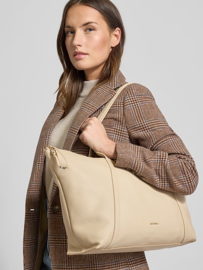 Kobiece modelka nosi torebkę z uchwytem Handtasche mit Tragehenkel Modell 'NORY' od Coccinelle, detal z logo, główna przegroda z zamkiem błyskawicznym, przegródka wewnętrzna zapinana na zamek, szerokość 50 cm, długość 30 cm, głębokość 14 cm, długość paska na ramię 60 cm, jasny beż, faktura skóry, całkowity widok z przodu.