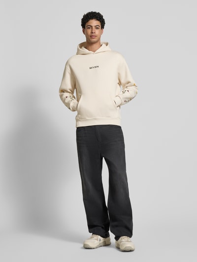Männermodel trägt Regular Fit Hoodie mit Logo-Stitching von REVIEW in Cremefarbe, florales Stitching an den Ärmeln, Känguru-Tasche, Ganzkörperansicht von vorne