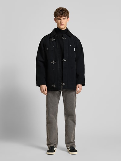 Männermodel trägt Oversized Jacke mit Logo-Patch Modell 'Adair' von Carhartt Work In Progress, Schwarz mit Cord-Besatz und Schnallen, Ganzkörperansicht von vorne.