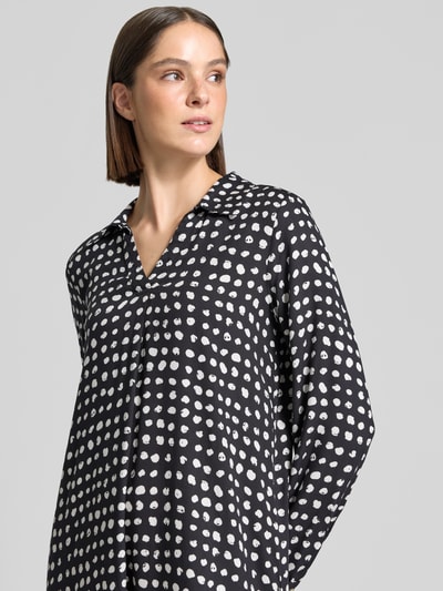 Frauenmodel trägt Knielanges Kleid aus reiner Viskose von Emily Van den Bergh mit weißem Punktemuster auf schwarzem Grund, Umlegekragen, langärmelig, relaxed Fit, Oberkörperansicht schräg von vorne, Rückenlänge 101 cm, Brustweite 51 cm, Ärmellänge 61 cm.