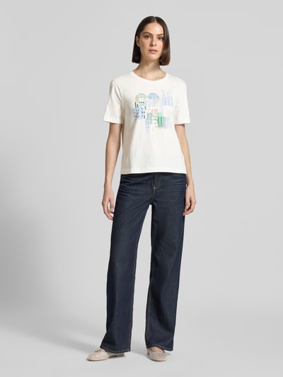 Frauenmodel trägt T-Shirt mit Motiv-Print und Pailletten von Marc Cain in Weiß, kombiniert mit Jeans, Baumwolle, Regular Fit, Ganzkörperansicht von vorne.