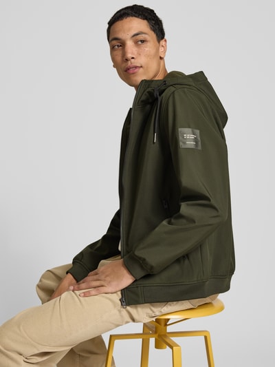 Männermodel trägt Regular Fit Jacke mit Logo-Detail von JACK & JONES in Olivgrün, Softshell, Kapuze, seitliche Taschen, Nahaufnahme Oberkörper. Rückenlänge, Schulterbreite, Brustweite und Ärmellänge bei Größe M im Kontext.