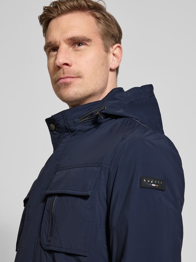 Mężczyzna nosi Regular Fit Jacke mit Stehkragen od Bugatti, granatowy materiał, aplikacja z logo, krój regular fit, kieszenie na piersi, kaptur, zbliżenie na górną część ciała