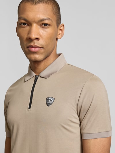 Męski model nosi Slim Fit Poloshirt aus Viskose-Mix von EA7 Emporio Armani, mieszanka wiskozy, detal z logo, kołnierzyk polo, elastyczne zakończenia rękawów, długość tyłu w rozmiarze M: 60 cm, jasnobeżowy kolor, tekstura gładka; zbliżenie na górną część ciała