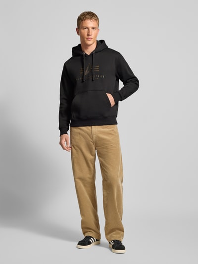 Männermodel trägt Hoodie mit Logo und Känguru-Tasche von Alpha Industries, Baumwoll-Mix, Regular-Fit, schwarze Farbe, Ganzkörperansicht von vorne.