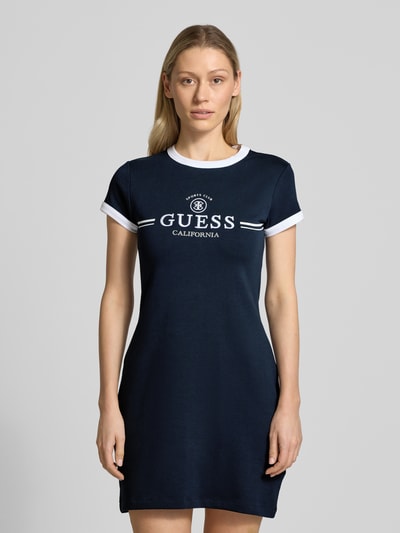Frauenmodel trägt Slim Fit Minikleid mit Logo-Stitching Modell 'Mila' von Guess, navy-blau mit weißen Details, elastischer Baumwoll-Mix, kurzärmelig, Rundhalsausschnitt, Ganzkörperansicht von vorne.