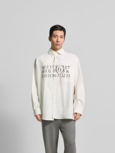 Männermodel trägt Relaxed Fit Freizeithemd mit Brusttasche von MM6 Maison Margiela in Creme mit schwarzem Aufdruck, Ganzkörperansicht von vorne, reine Baumwolle, Kentkragen, durchgehende Knopfleiste, lockerer Schnitt.