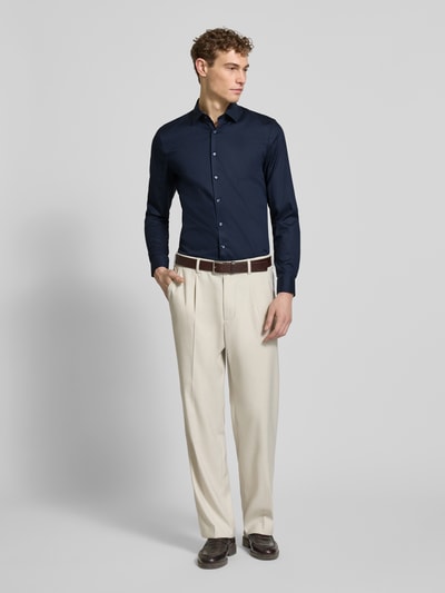 Męski model nosi Slim Fit Businesshemd mit Kentkragen von OLYMP No. Six, elastyczna mieszanka bawełny, krój slim fit, kołnierzyk kent, pełna listwa guzikowa, długi rękaw, sportowe mankiety, długość tyłu w rozmiarze 40: 81 cm, szerokość ramion w rozmiarze 40: 43 cm, długość rękawa w rozmiarze 40: 67 cm, granatowa góra, jasne spodnie, widok całej sylwetki z przodu.
