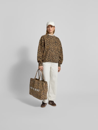 Frauenmodel trägt Sweatshirt mit Allover-Animal-Print von Anine Bing, Relaxed-Fit, Logo-Stitching, Rundhalsausschnitt, elastische Abschlüsse, cremefarbene Hose, braune Schuhe, große Tasche, Beige- und Brauntöne, weiche Textur, Ganzkörperansicht von vorne.