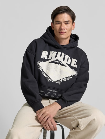 Männermodel trägt Sweatshirt mit großem Frontprint von RHUDE in Schwarz mit weißem Motiv-Print, Oversized Longline-Passform, Sweatmaterial, Nahaufnahme Oberkörper.