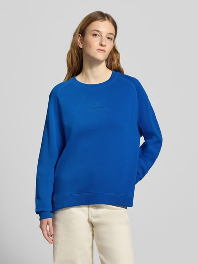 Modelka kobieca nosi Regular Fit Sweatshirt aus reiner Baumwolle od Marc O'Polo Denim, kolor niebieski, bawełniany materiał, napis z przodu, krój regular fit, długie rękawy, gładka faktura, okrągły dekolt, widok całej sylwetki z przodu.