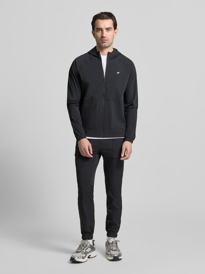 Männermodel trägt zwarte 'Trainingsjacke mit Kapuze Modell THE ONE JACKET' van Fabletics met reflecterend logo, rechte pasvorm, doorlopende rits en elastische boorden, ganzkörperansicht von vorne.