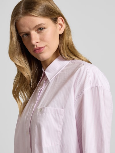 Frauenmodel trägt Oversized Hemdbluse mit Brusttasche von Guess Jeans in zartem Rosa mit feinen Streifen, Reine Baumwolle, Logo-Patch, Oversized-Fit, Hemdblusenkragen, Langärmeliges Design, durchgängige Knopfleiste, abgerundeter Saum und verlängerter Rücken, Nahaufnahme Oberkörper.