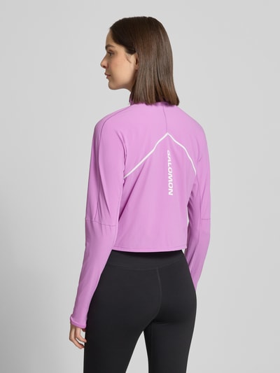 Modelka nosi bluzkę z długim rękawem 'Langarmshirt im Cropped-Design mit Stehkragen' od Salomon w kolorze jasnoróżowym, taliowany krój, krótki fason, stójka z ochroną podbródka, half-zip, tunel w pasie, rękawy długie, długość tyłu w rozmiarze S: 47 cm, widok sylwetki z tyłu.