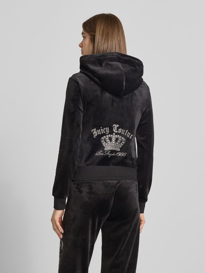 Kobiece modelka nosi Sweatjacke mit Ziersteinbesatz Modell 'GOTHIC CROWN ROBERTSON' von Juicy Couture, czarny materiał z połyskiem, krój slim fit, kaptur z troczkiem, dwukierunkowy zamek, widoczny napis i aplikacja z tyłu, widok całej sylwetki z tyłu.