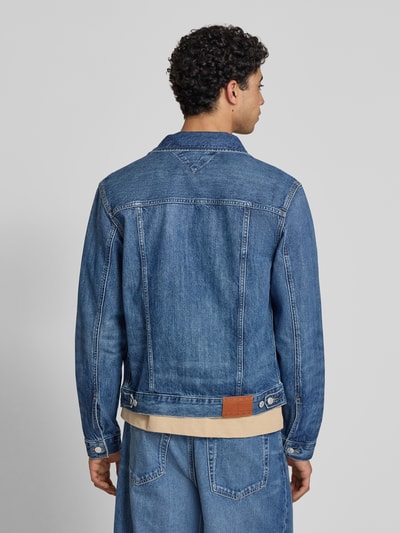 Männermodel trägt Regular Fit Jeansjacke aus reiner Baumwolle Modell 'RYAN' von Tommy Jeans w kolorze niebieski denim, splot twill, długi rękaw, naszywka z logo, krój regular fit, wykładany kołnierz, listwa guzikowa, nity, kieszenie boczne i na piersi, tylny pas, tylna perspektywa pełnej sylwetki.