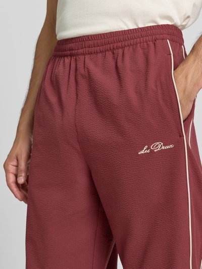 Männermodel trägt Trackpants mit Logo-Stitching Modell 'Seersucker' von Les Deux in Dunkelrot mit Strukturmuster und Kontraststreifen, Nahaufnahme Oberkörper. Relaxed Fit, elastischer Bund mit Tunnelzug, Eingrifftaschen, Innenbeinlänge bei Größe M 78 cm.