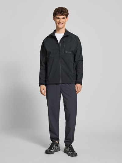 Männermodel trägt Softshell-Jacke mit Reißverschlusstaschen Modell 'ELITE' von HALO in Schwarz, Regular-Fit, Stehkragen, Label-Print, Ganzkörperansicht von vorne. Rückenlänge 71 cm, Schulterbreite 55 cm, Brustweite 60 cm, Ärmellänge 63 cm.