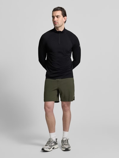 Männermodel trägt Slim Fit Longsleeve mit Logo-Print von Fabletics in Schwarz, Troyer-Kragen, Kinnschutz, Ganzkörperansicht von vorne.