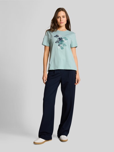 Frauenmodel trägt Regular Fit T-Shirt aus reiner Baumwolle von Tom Tailor in Mint mit floralem Motiv-Print und kurzem Arm, kombiniert zu dunkelblauer Hose, unifarbene Optik, Jerseytextur, Ganzkörperansicht von vorne.