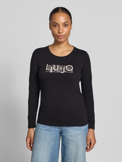 Frauenmodel trägt Langarmshirt aus Baumwoll-Mix von Liu Jo, schmale Passform, Rundhalsausschnitt, schwarzer Stoff, Label-Print, Ziersteine, Ganzkörperansicht von vorne.