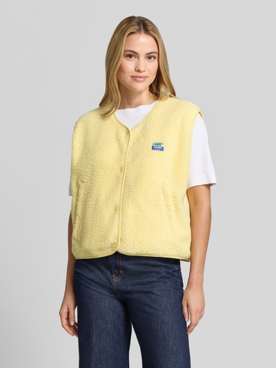 Frauenmodel trägt Relaxed Fit Weste aus Teddyfell Modell 'Hoktown' von American Vintage, gelb, Knopfleiste, Logo-Patch, Nahaufnahme Oberkörper. Damen Weste, Rückenlänge 56 cm, Schulterbreite 43 cm, Brustweite 63 cm.