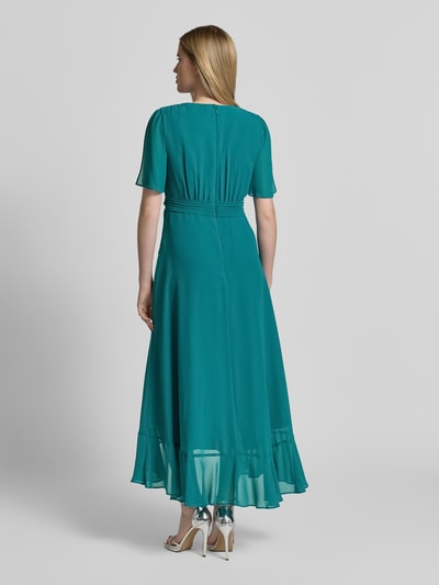 Frauenmodel trägt Cocktailkleid in Wickel-Optik mit Volantsaum von Christian Berg Cocktail in Türkis, fließender Chiffon, tailliert, Ganzkörperansicht Rückansicht.