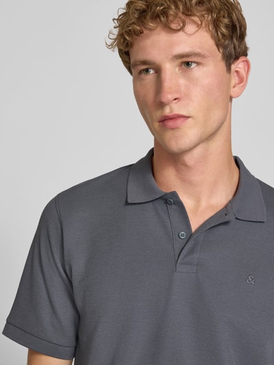 Mężczyzna nosi koszulkę polo Slim Fit Poloshirt mit Label-Stitching Modell 'AUSTIN' von JACK & JONES, mieszanka materiałów z bawełną, kołnierzyk polo, wyhaftowane logo, długość tyłu w rozmiarze M: 70 cm, szara faktura, prosty krój, zbliżenie na górną część ciała.