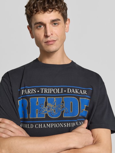 Männermodel trägt Regular Fit T-Shirt aus Baumwolle von RHUDE mit Label-Print, schwarzer Stoff, blaue und goldene Schrift, Nahaufnahme Oberkörper.