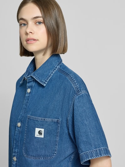 Vrouwenmodel draagt Relaxed Fit overhemdblouse in denimlook model 'Lovilia' van Carhartt Work In Progress, puur katoen, borstzak met logopatch, korte mouw, overhemdkraag, knoopsluiting, detailopname bovenlichaam.