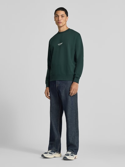 Männermodel trägt Regular Fit Sweatshirt aus Baumwoll-Mix Modell 'Soho' von JACK & JONES in Dunkelgrün, Logo-Print, Ganzkörperansicht von vorne. Rückenlänge 65 cm, Ärmellänge 66 cm, Rundhalsausschnitt, elastische Abschlüsse.